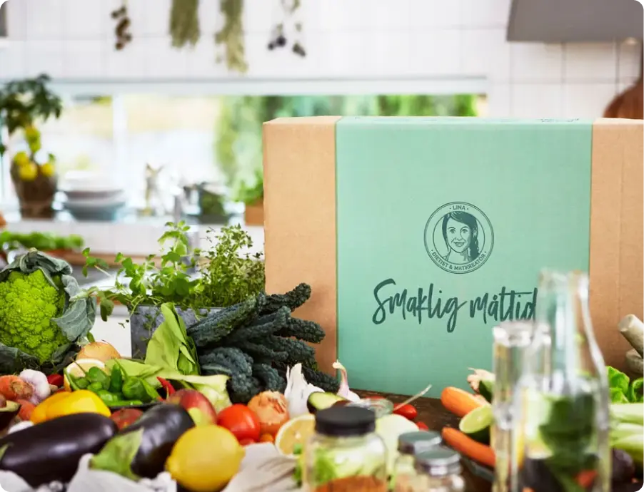 Kitchen counter with plenty of vegetables and a cardboard box with a brand logo and the text Smaklig måltid