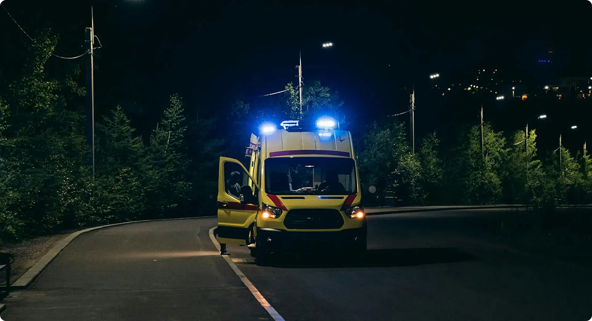 Ambulance with blue lights parked on a road surrounded by trees at night